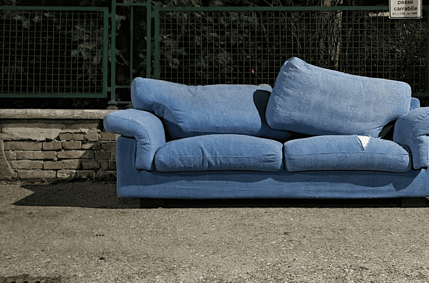 Blue upholstered sofa with three seat cushions and three backrest cushions sits outdoors, discarded on a sidewalk near a metal fence and brick wall.