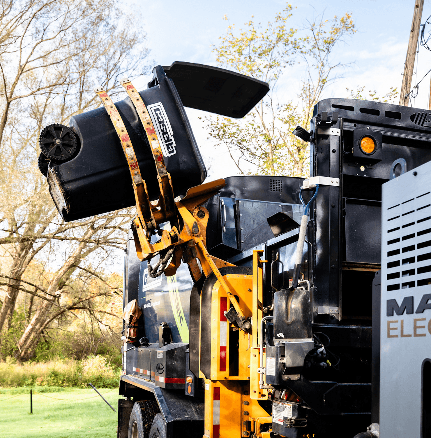 A Casella automated side-load truck uses its mechanical arm to lift a garbage cart and empty its contents into the truck's hopper.