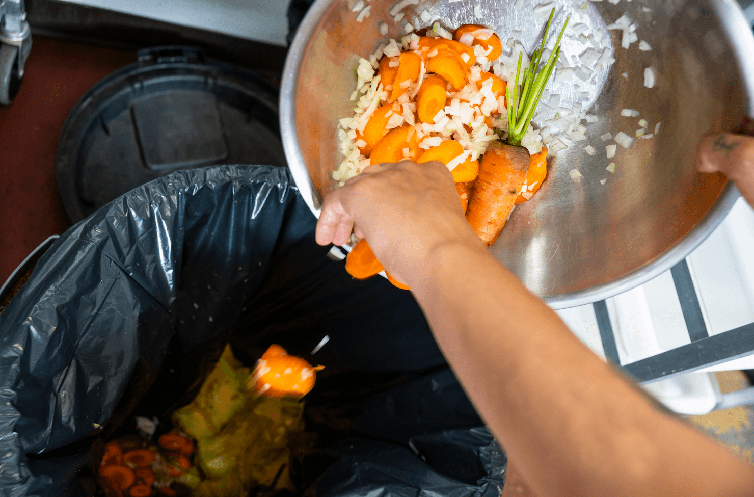 Person discarding food scraps like carrots and onions into a designated food waste bin.