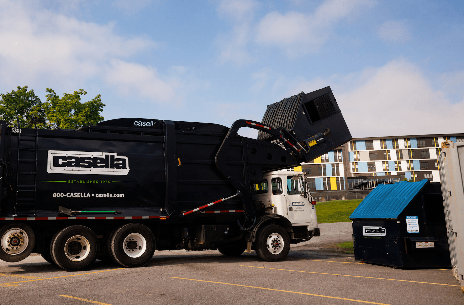 Casella front-load truck servicing a commercial dumpster in a grassy, rural area.